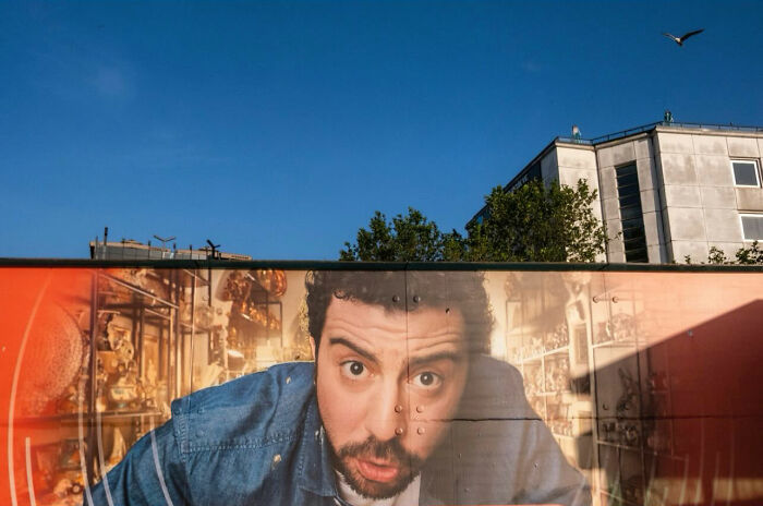 Street photograph of a surprised man in a blue shirt with clear timing, featuring urban buildings and a bird in the sky above.