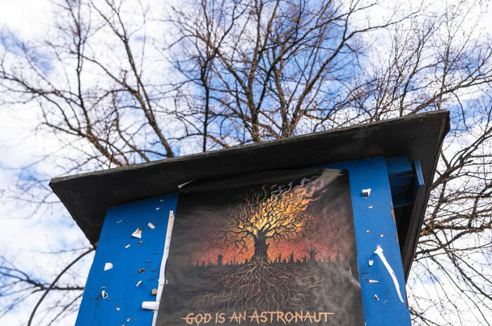 Street photograph of a poster with a fiery tree and text God Is An Astronaut under leafless trees and a partly cloudy sky.