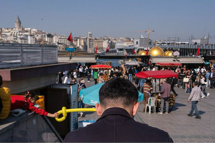 Man holding yellow balloon sword, busy street scene with vendors and people, capturing entertaining street photographs timing.