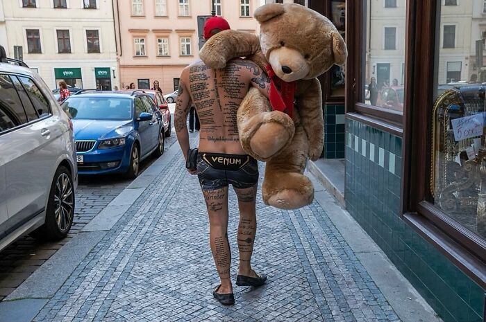 Man carrying large teddy bear on street with tattooed back and timing caught in entertaining street photograph.