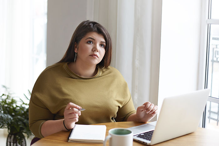 Overweight woman working at a laptop, appearing thoughtful in a bright office setting with notebook and coffee mug.