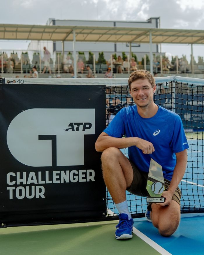 Young male tennis player kneeling on court holding trophy next to ATP Challenger Tour banner during match event Young male tennis player kneeling on court holding trophy next to ATP Challenger Tour banner during match event