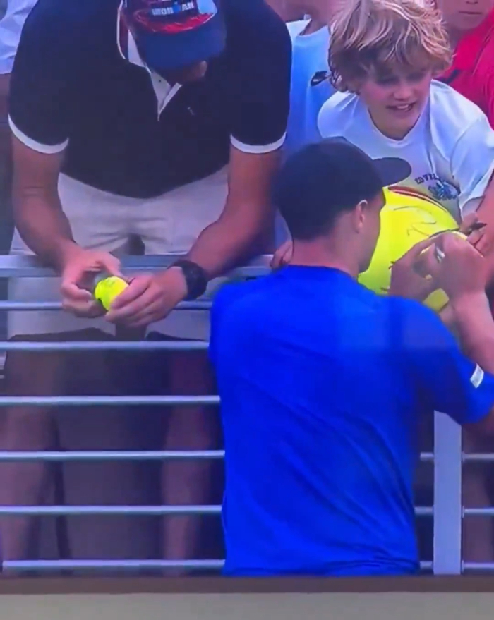 Man in blue shirt signing tennis balls for fans at a US Open tennis event amid viral CEO cap incident. Man in blue shirt signing tennis balls for fans at a US Open tennis event amid viral CEO cap incident.
