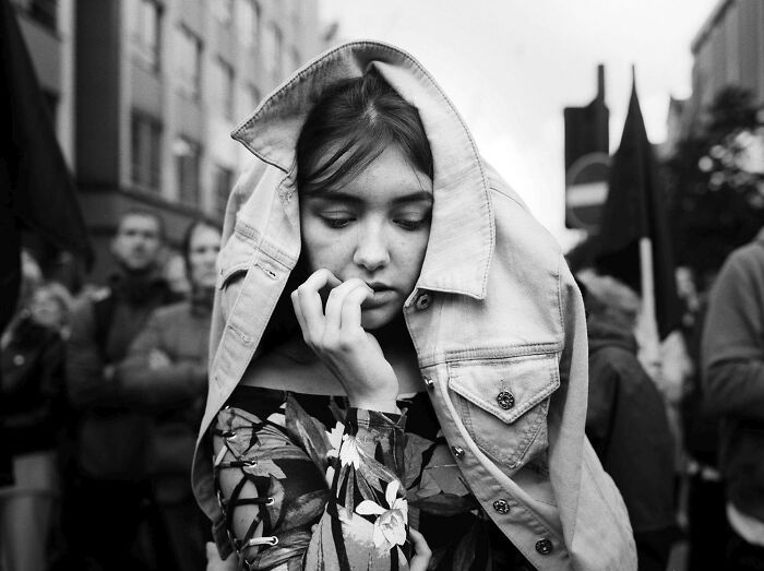 Black and white analog photo of a pensive woman with a jacket over her head, captured at a street event.