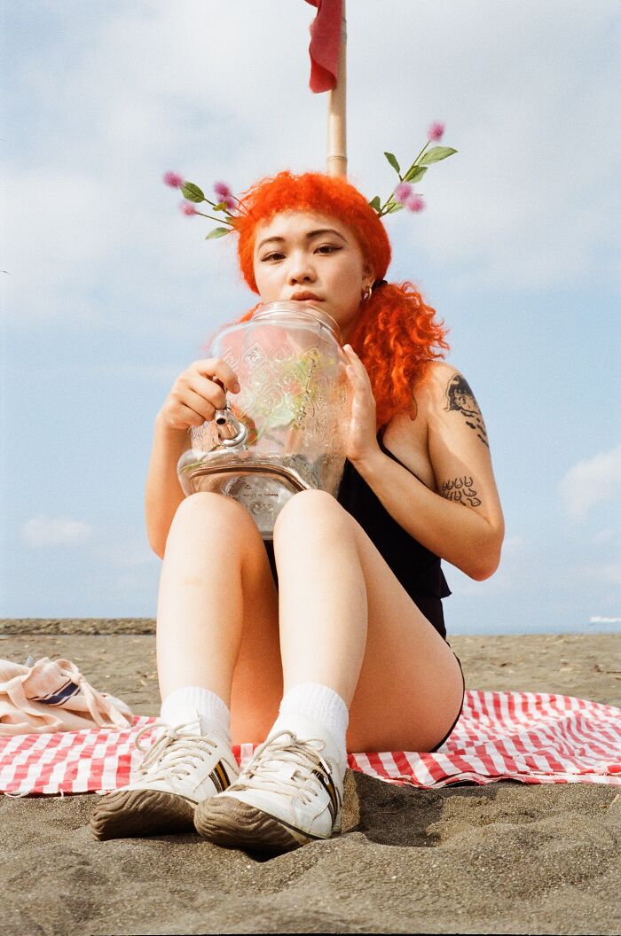 Young woman with bright orange hair sitting on the beach holding a glass jar, captured in an analog photo style.