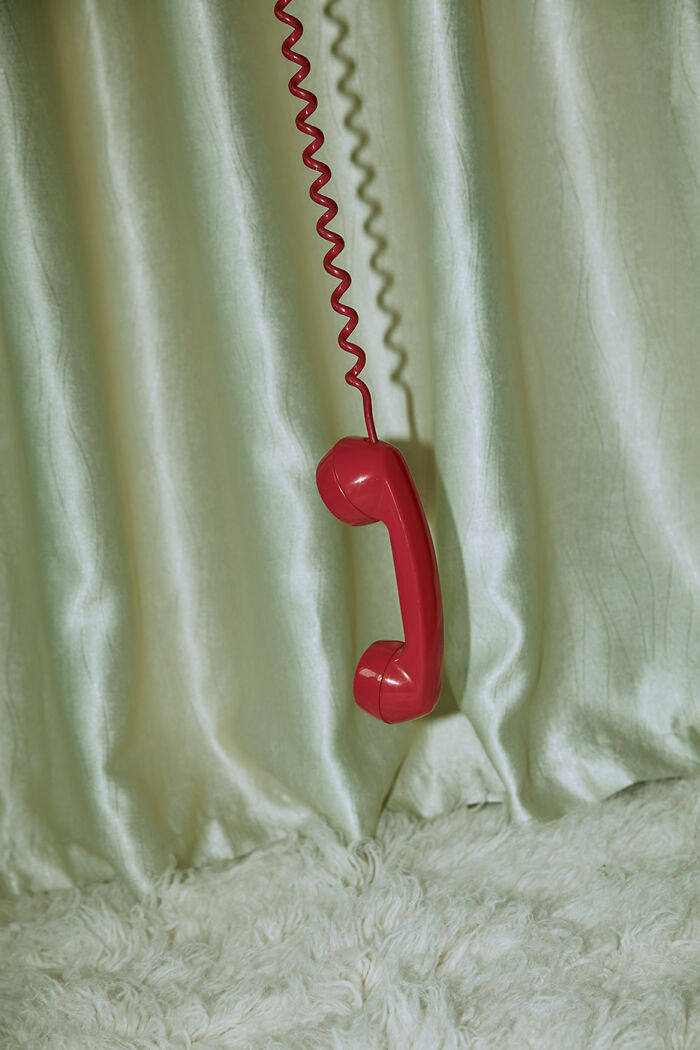 Red telephone handset suspended against a pale curtain with a simple textured rug, showcasing minimalism style.