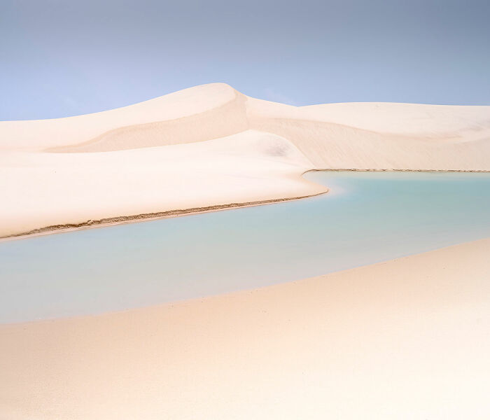 Minimalism landscape with smooth sand dunes and a calm blue water body under a clear sky in a serene setting.