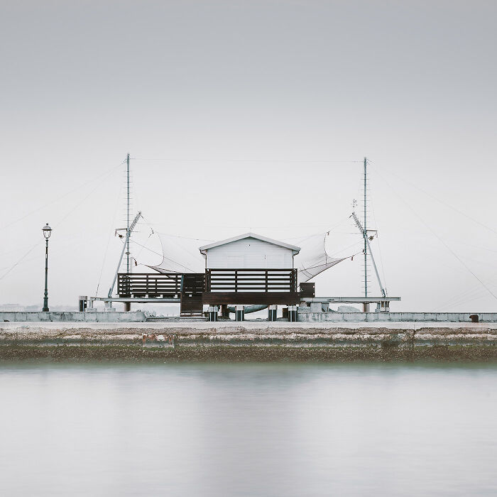 Minimalist photo of a simple wooden structure by calm water under a pale, overcast sky.