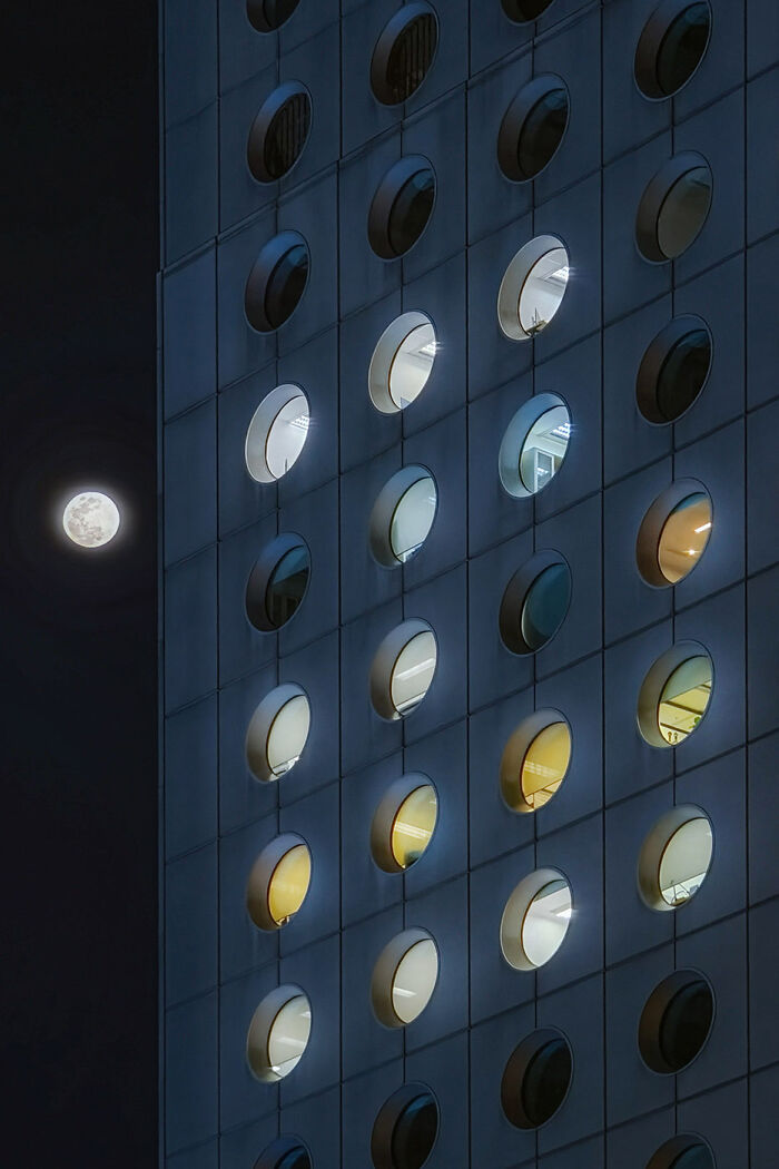 Night view of a minimalist building facade with circular windows lit from inside and a full moon in the dark sky.