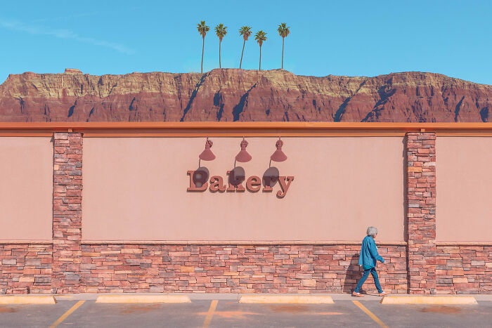 Person walking past a bakery wall with minimalist design, clear blue sky, and a row of palm trees against rocky cliffs.