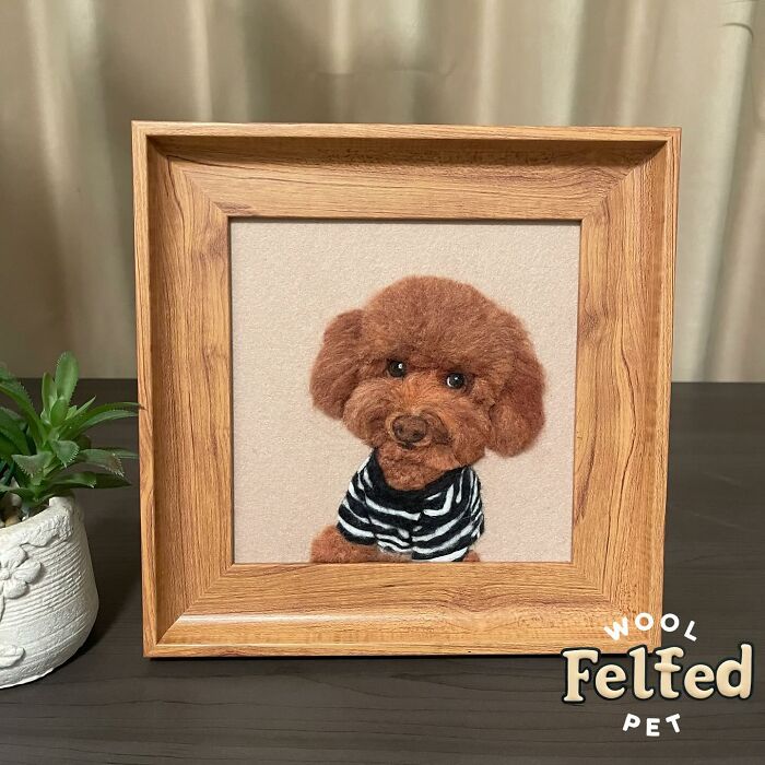 Realistic wool felt pet portrait of a brown dog wearing a striped shirt in a wooden frame on a dark table.