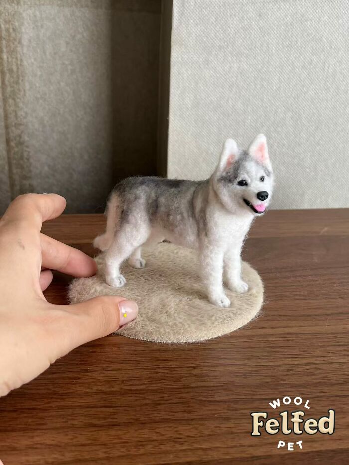 Hand holding a realistic wool felt pet dog sculpture on a round wool felt base, showing detailed craftwork.