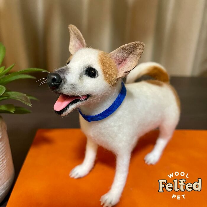 Realistic wool felt pet dog with brown patches and blue collar displayed on orange surface next to green plant.