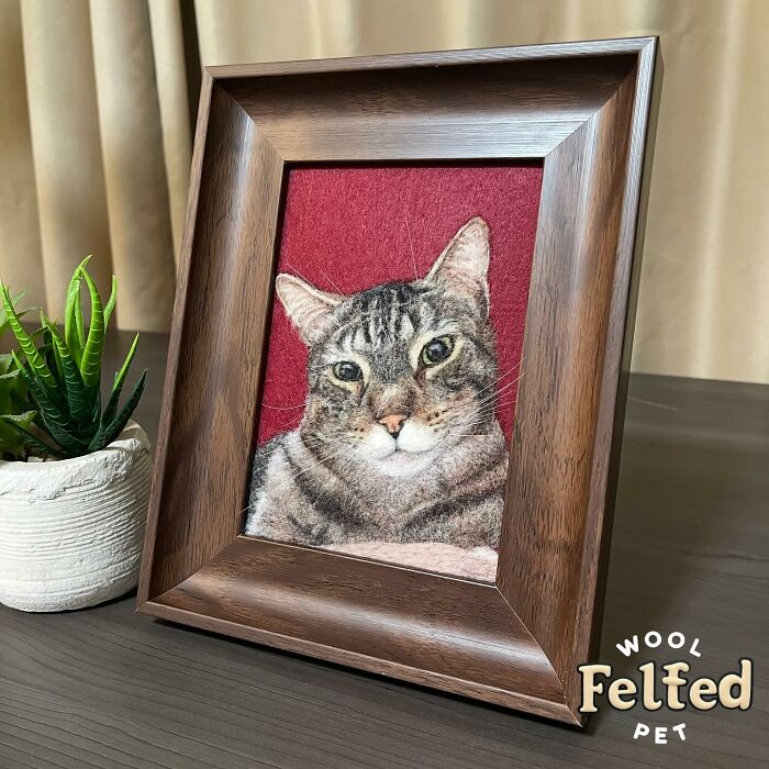 Framed wool felt pet portrait of a realistic tabby cat on a red background, displayed on a wooden table.