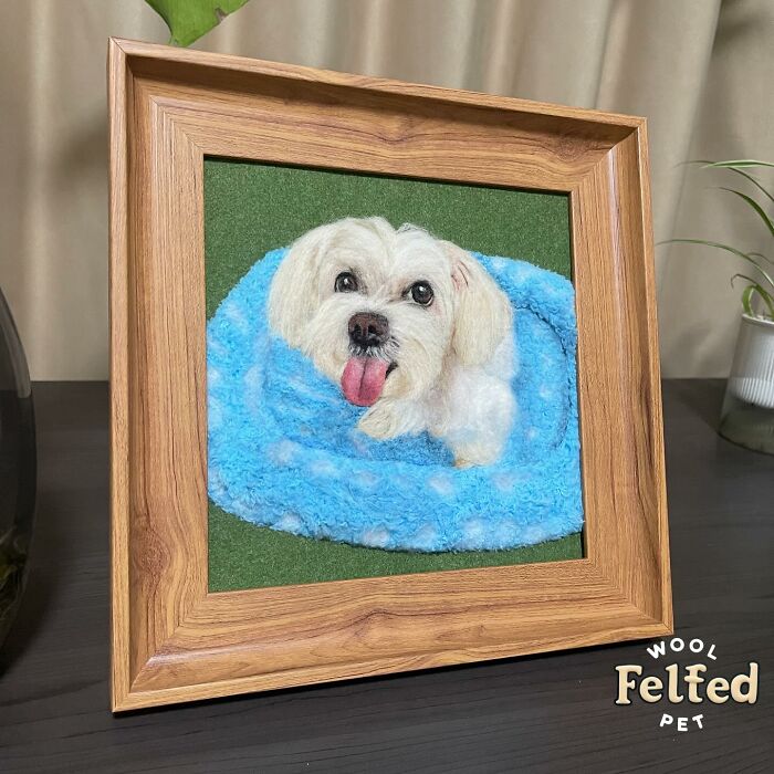 Realistic wool felt pet artwork of a small white dog lying on a blue blanket, framed in wood on a table.