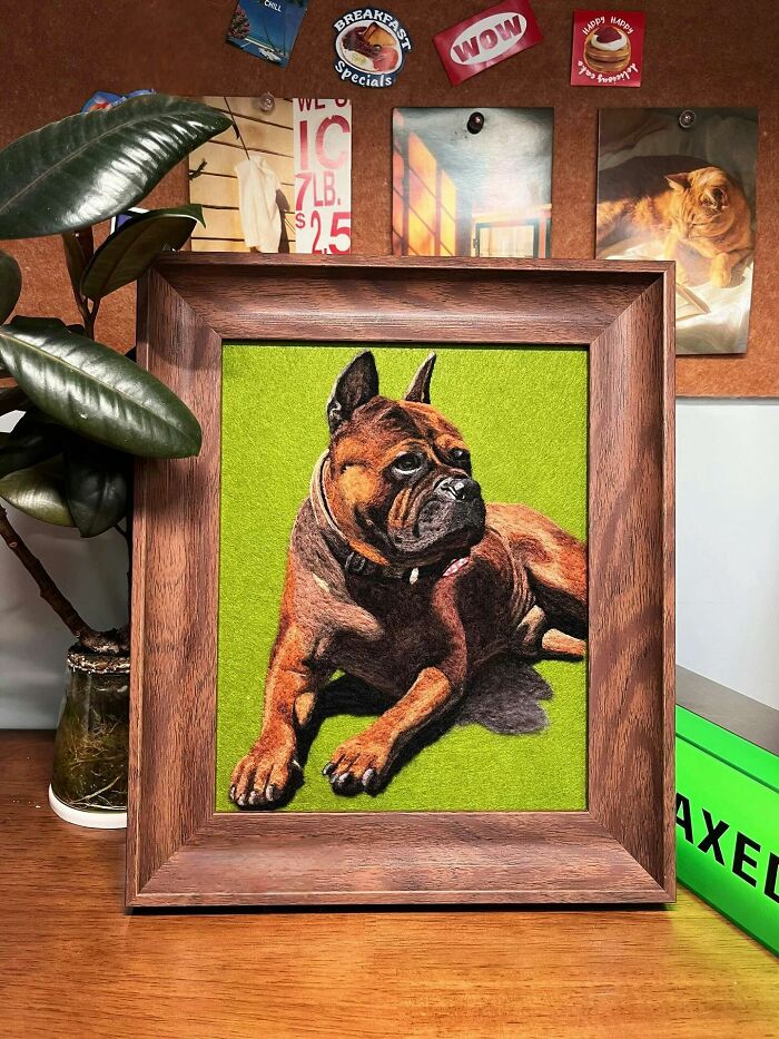Framed wool felt pet portrait of a realistic dog on a green background, displayed on a wooden surface.