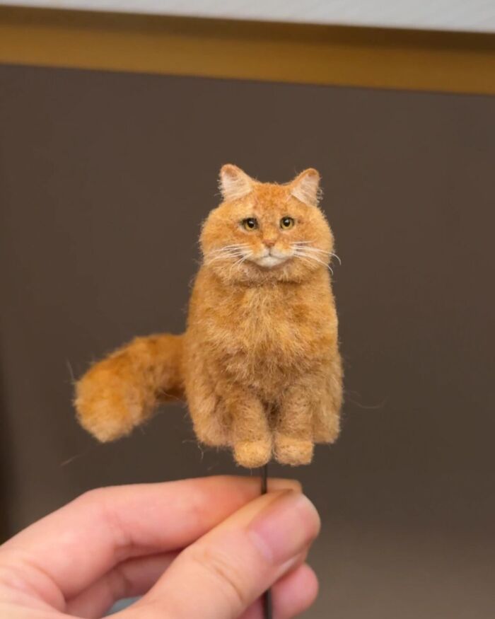 Tiny wool felt pet resembling a realistic orange cat held between fingers, showcasing detailed fur and lifelike features.