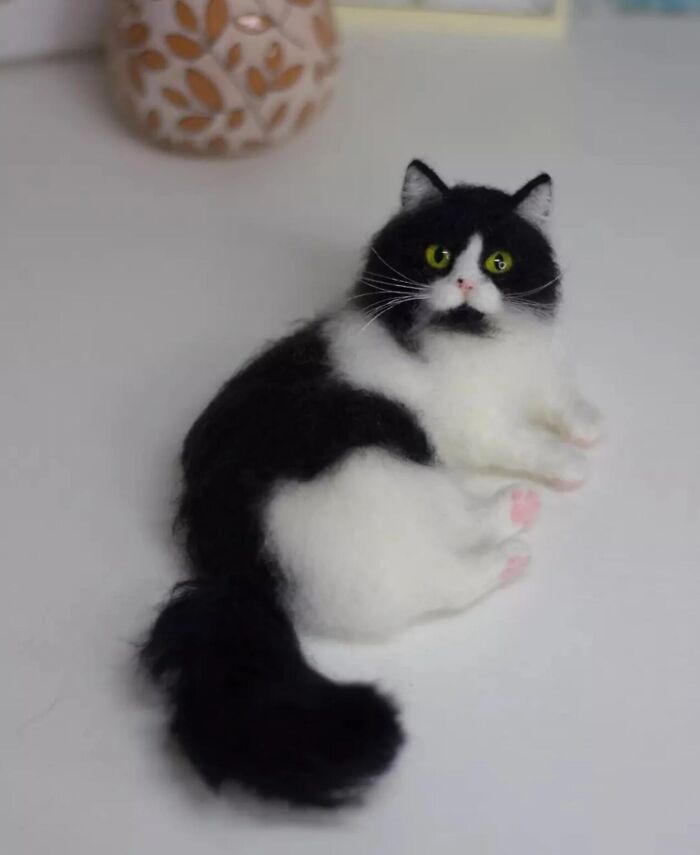 Black and white wool felt pet cat with green eyes, crafted to look incredibly realistic on a white surface.