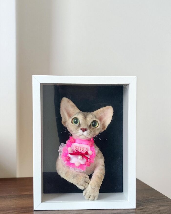 Realistic wool felt pet cat sculpture with big eyes and pink decorative collar displayed in white frame.