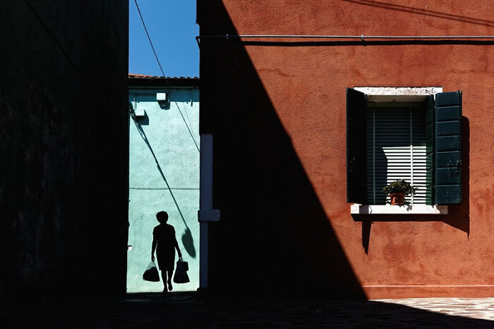 Silhouette of a person walking through an alley with contrasting shadows and vibrant building colors in a stunning photo.