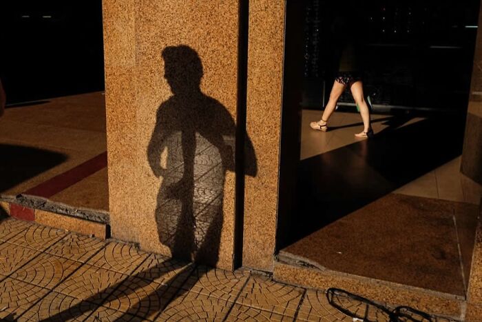 Shadow of a person cast on a textured wall with a passerby walking in a brightly lit urban setting, stunning photos.