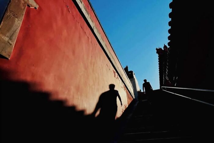 Silhouettes of people walking up stairs beside a red wall under clear blue sky in a stunning photo from around the world.