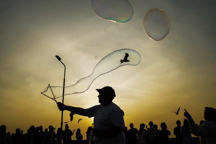 Silhouetted person creating large soap bubbles at sunset among a crowd in a stunning travel photo from around the world.