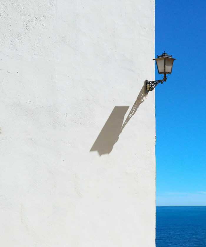 Minimalist photo of a wall lamp casting a shadow on a white wall with a bright blue sky and ocean in the background.