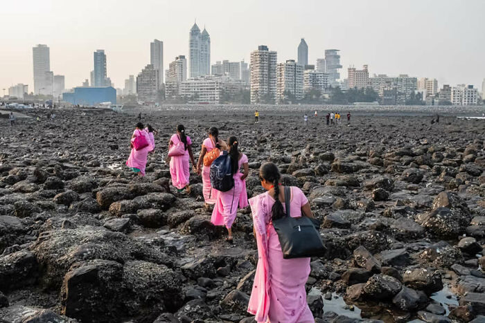 Women in pink saris walking on rocky shore with city skyline in background, showcasing stunning photos from around the world.