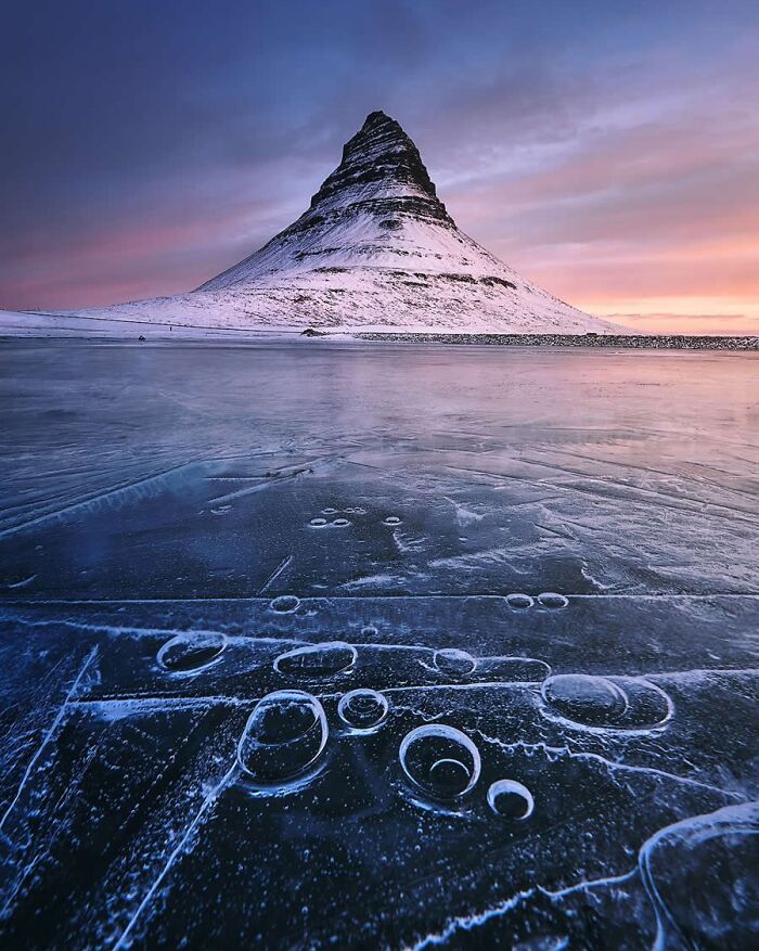 Snow-covered mountain during sunset with frozen lake in foreground, featured on Instagram page collecting stunning photos.