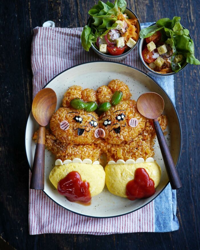 Creative Japanese kids meal featuring cute rice characters with omelet feet, served with fresh salad and wooden spoons on a plate.