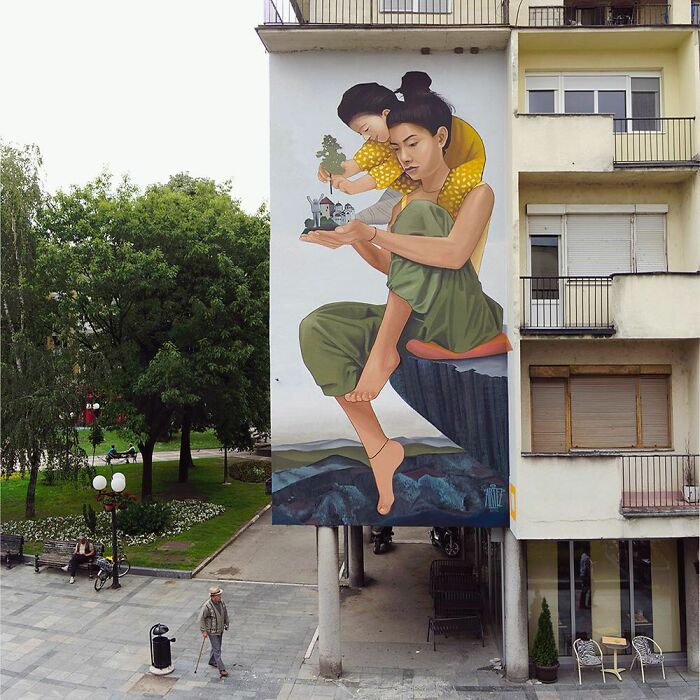 Large colorful mural by Artez showing a woman and child holding a miniature city, brightening an urban building facade.