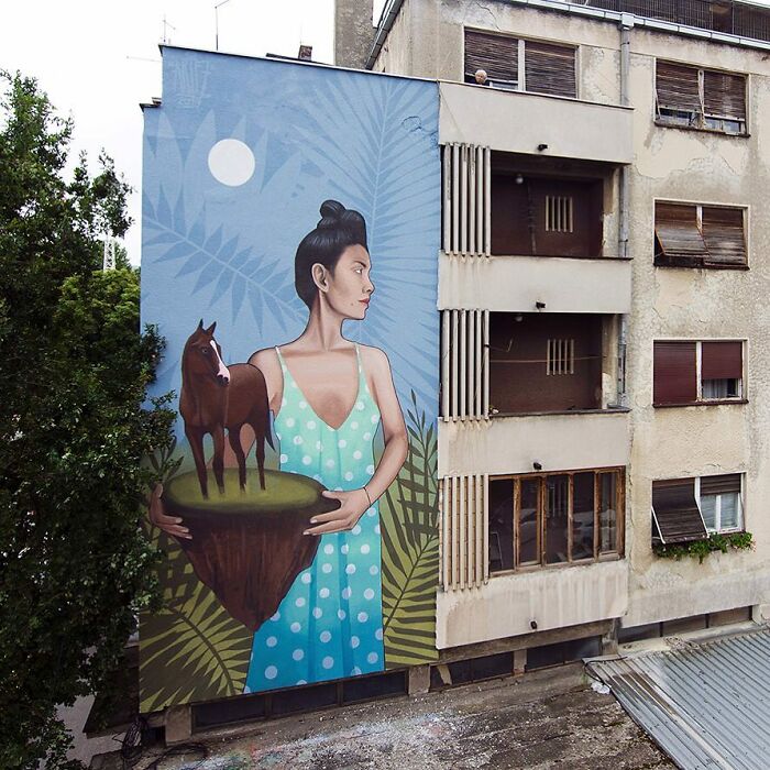 Large colorful mural by Artez of a woman in a blue polka dot dress holding a floating island with a horse on it in an urban setting.