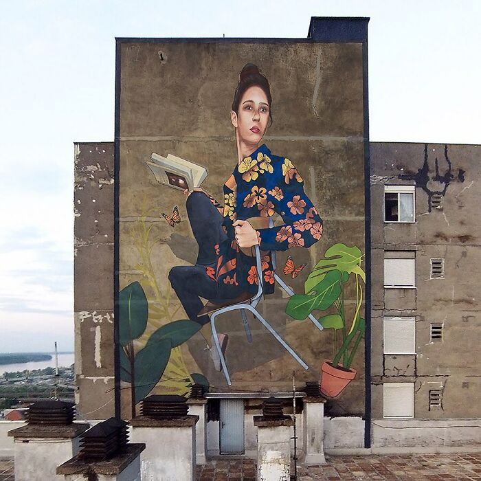 Colorful mural by Artez depicting a woman reading with butterflies and plants on a large city building wall.