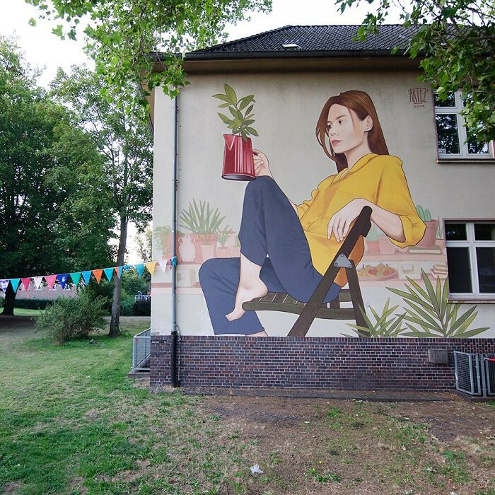 Large colorful mural by Artez of a woman in a yellow shirt holding a red watering can, bringing city walls to life.