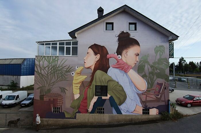 Large colorful mural by Artez on a city building wall featuring two women with hand puppets and indoor plants.