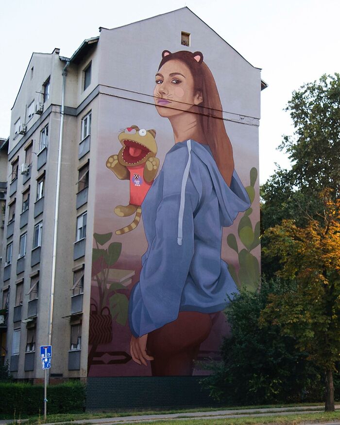 Large colorful mural on building features a girl in a blue hoodie with a cartoon cat puppet, showcasing vibrant city street art.