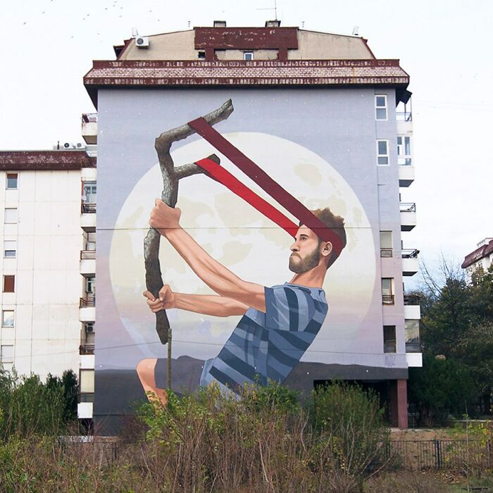 Large colorful mural by Artez on a building depicting a man with a slingshot against a full moon background.
