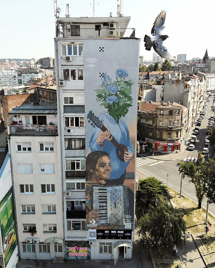 Large colorful mural by Artez on a city building, featuring a person with flowers for a head and another playing an accordion.