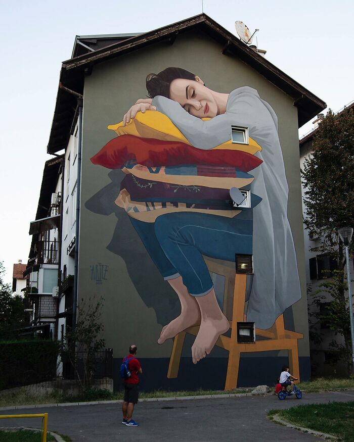 Large colorful mural by Artez of a woman sleeping on stacked pillows while sitting on a chair on a building wall.