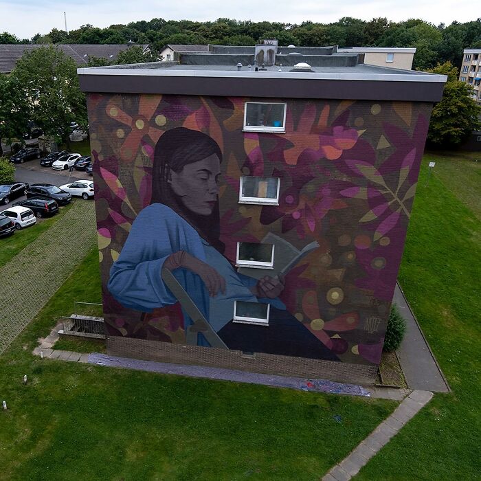 Large colorful mural by Artez of a woman reading, painted on a city building wall surrounded by greenery and parked cars.