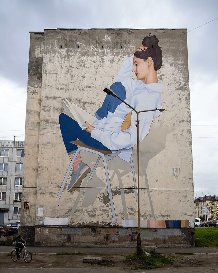 Large colorful mural by Artez on a city building showing a woman reading while sitting thoughtfully on a chair.