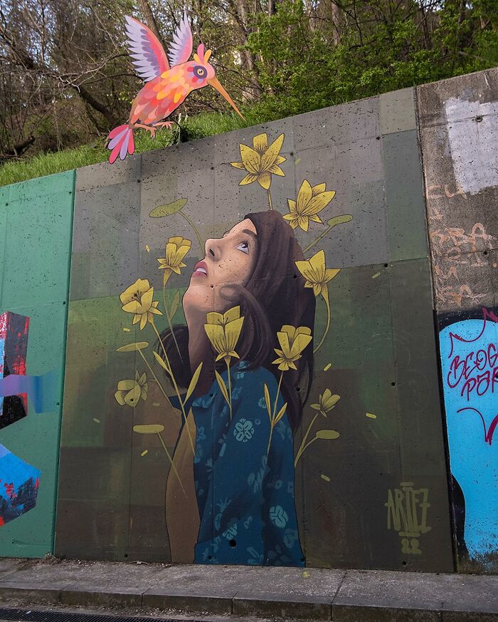 Colorful mural by Artez depicting a woman surrounded by yellow flowers looking up at a vibrant bird on a city wall.