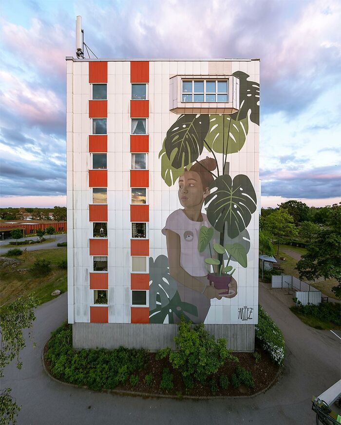 Large colorful mural by Artez of a girl holding a potted plant on the side of a city building at sunset.