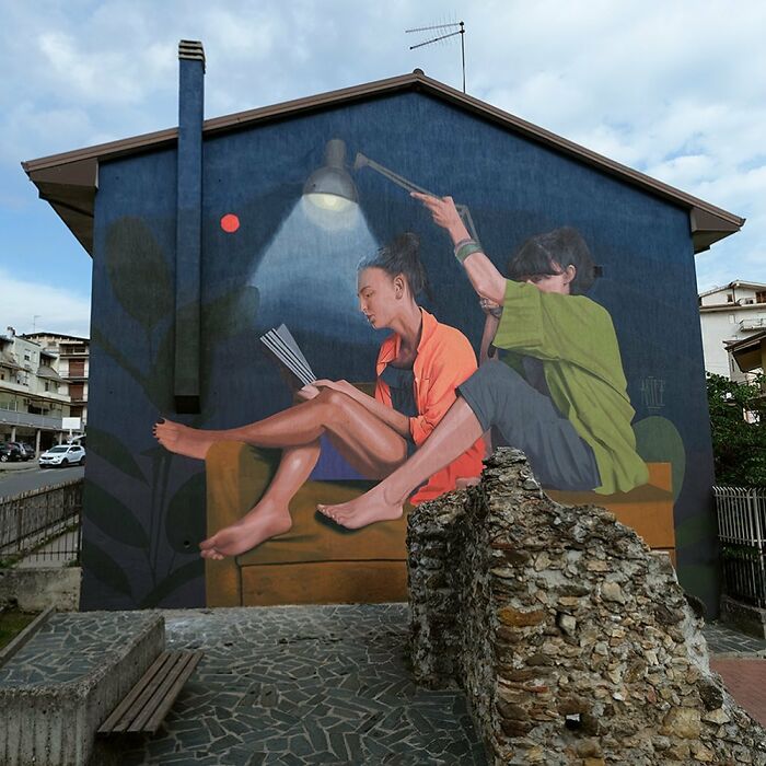 Colorful murals by Artez showing two people reading and interacting under a lamp on the side of a building in a city.