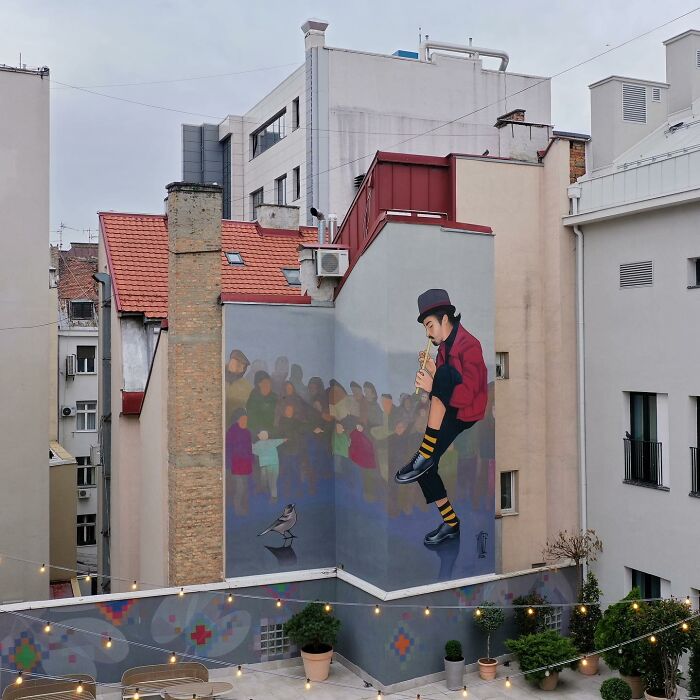 Colorful mural by Artez on a city building wall showing a musician playing flute with a crowd and bird nearby.