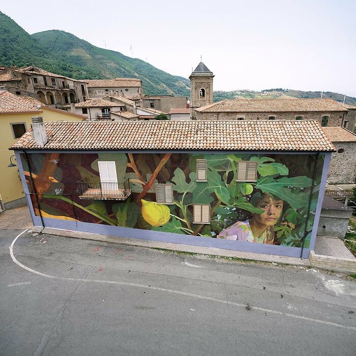 Colorful mural by Artez featuring a woman and large green leaves on a building wall in a small town setting.