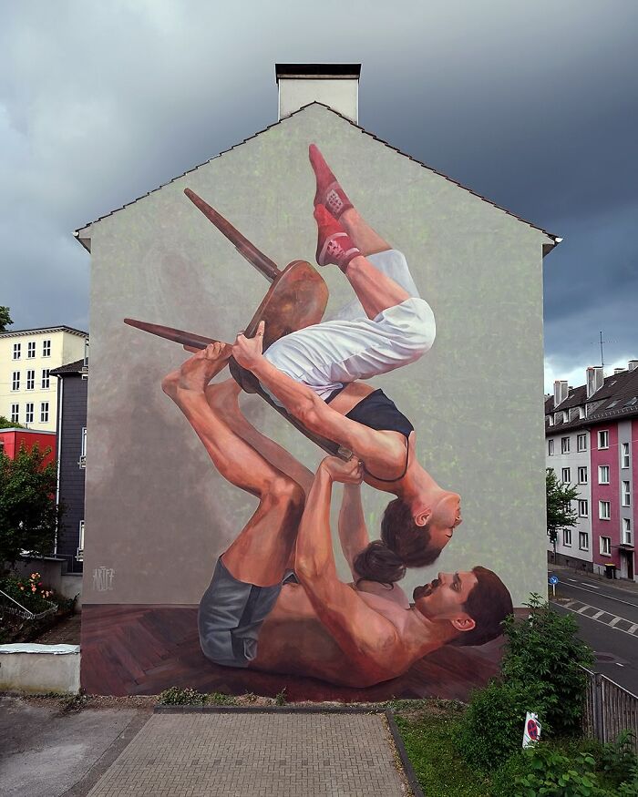 Large colorful mural by Artez depicting two people performing an acrobatic pose on a city building wall.