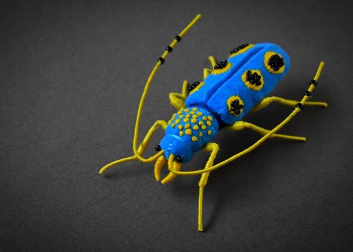 Bright blue and yellow felted artwork of an insect with detailed legs and antennae on a dark background.