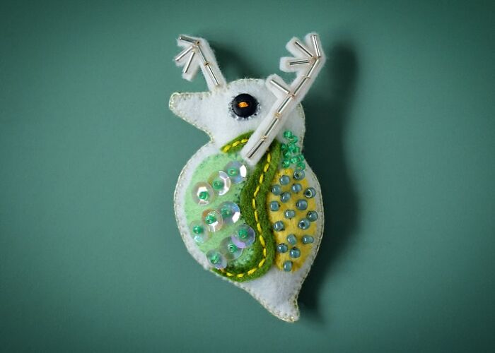 Small felted artwork of a whimsical creature decorated with beads and stitched details on a green background.